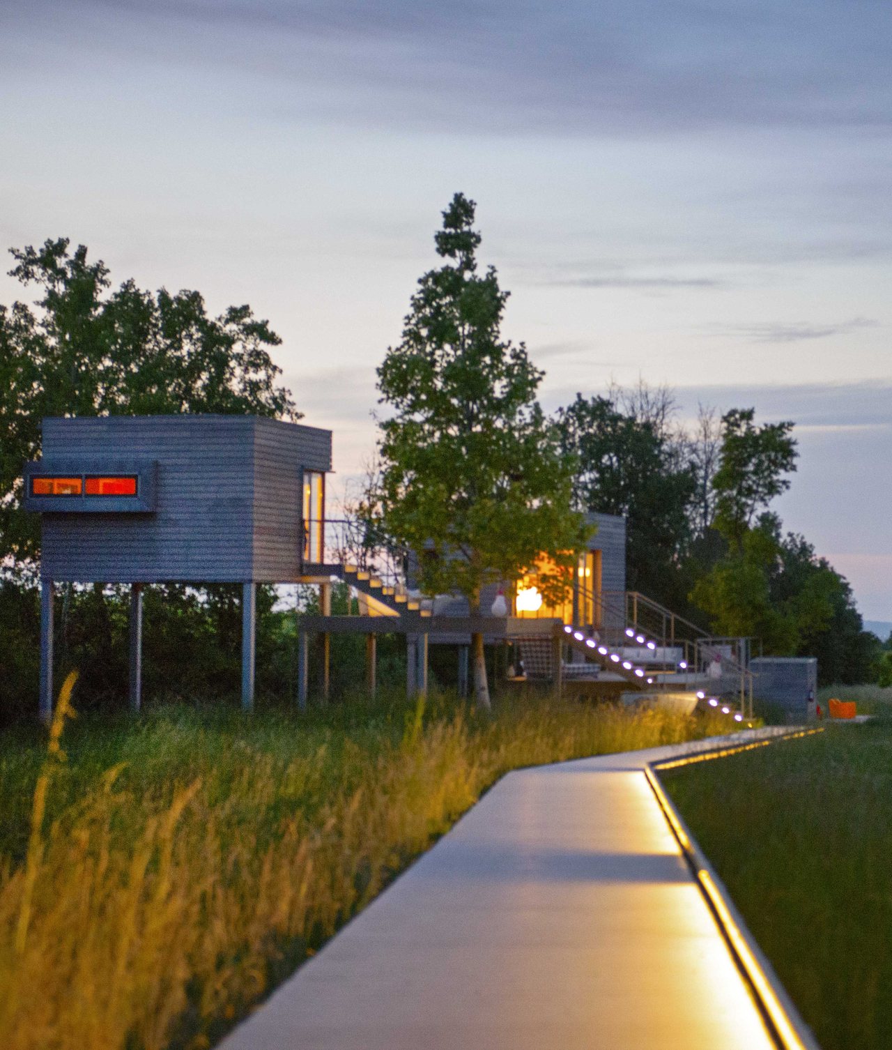 Case in legno sollevate su pilastri al tramonto, con le finestre accese e una passerella illuminata nel prato